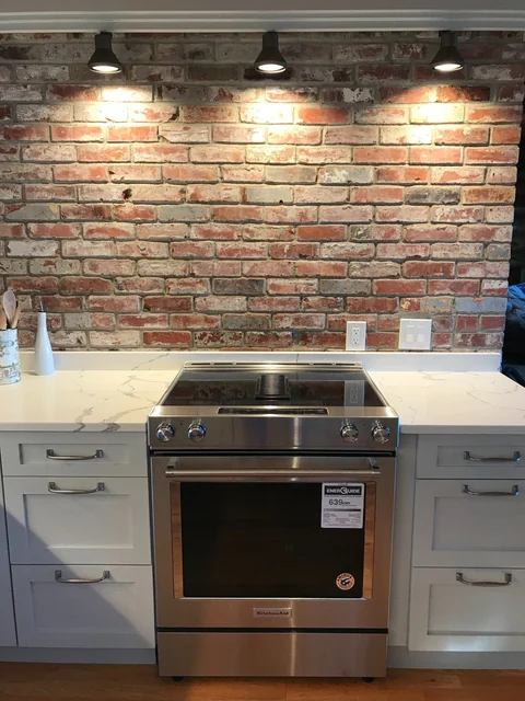 Kitchen range wall with white base cabinets, quartz counters and exposed brick backsplash